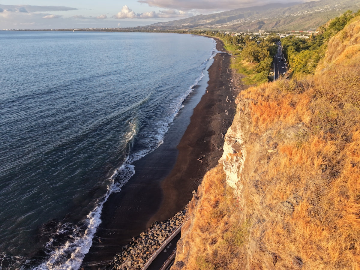 Coastal view with dark beach, ocean, and sunlit cliffs.
